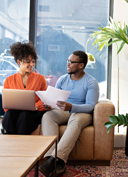 A man and women sat on a sofa with a laptop and papers talking in an office