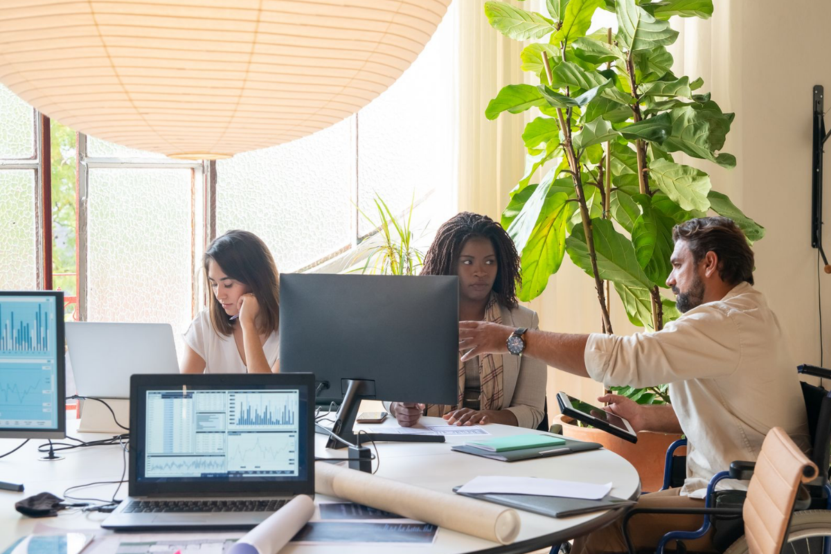 Three colleagues work around a shared office table, with charts on a laptop and monitor as one person holds a tablet and gestures toward a colleague’s screen.