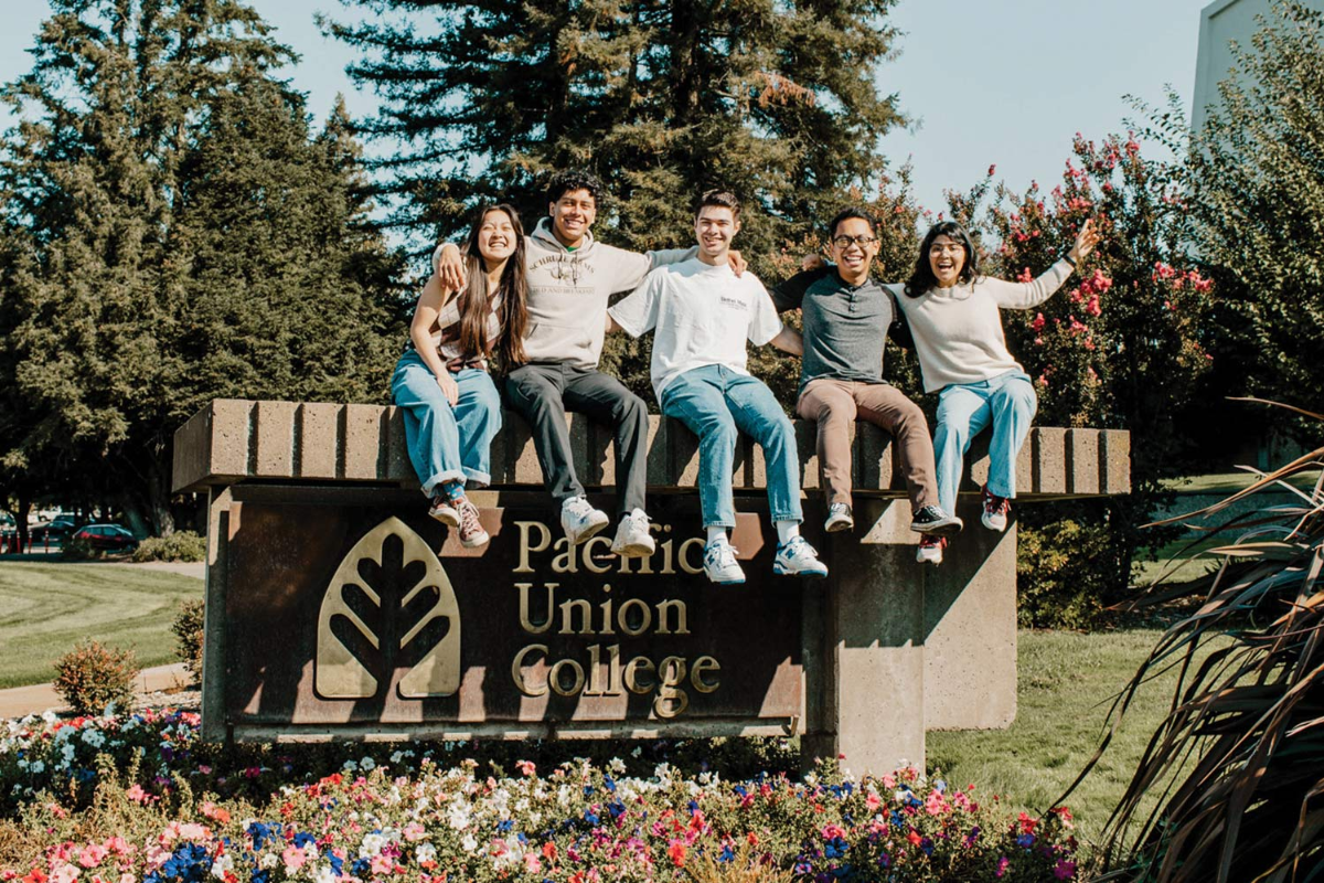 Photo of the sign of Pacific Union College with students sitting on top of it smiling and cheering.