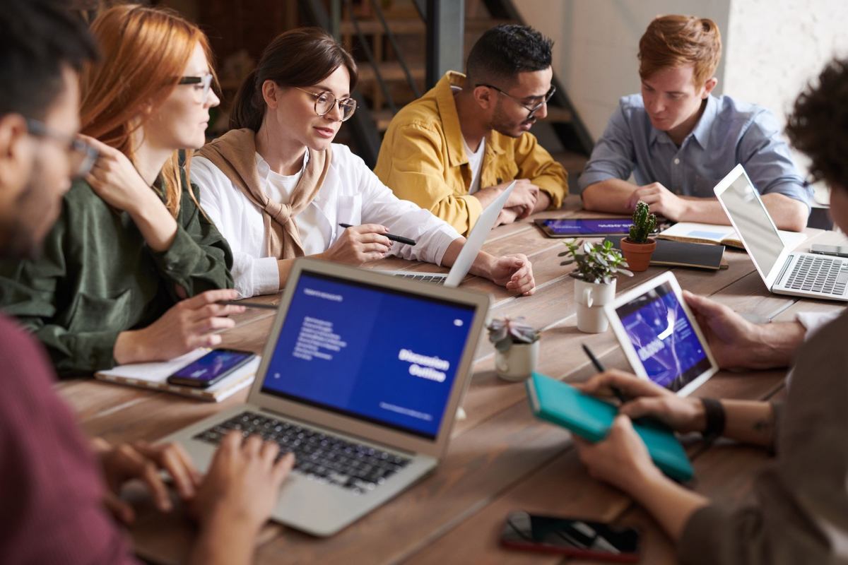 A team collaborates around a wooden table using laptops and tablets displaying digital content.