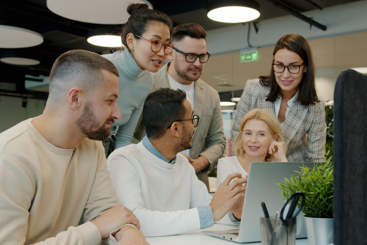 Six colleagues gather around a laptop in a modern office, with a seated man gesturing toward the screen as the group looks on. They seem to be discussing work presented in the laptop screen.