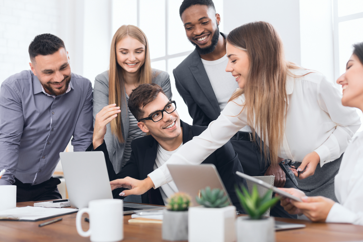 Six smiling colleagues collaborate around two laptops at an office table, with one person reaching across to point at a screen.