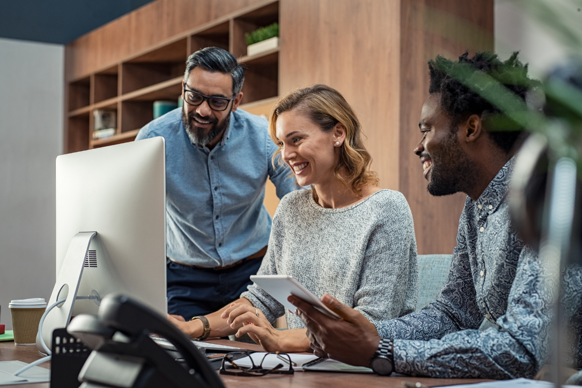 Three colleagues smile as they review content on a desktop computer, with one holding a tablet, in a modern office.