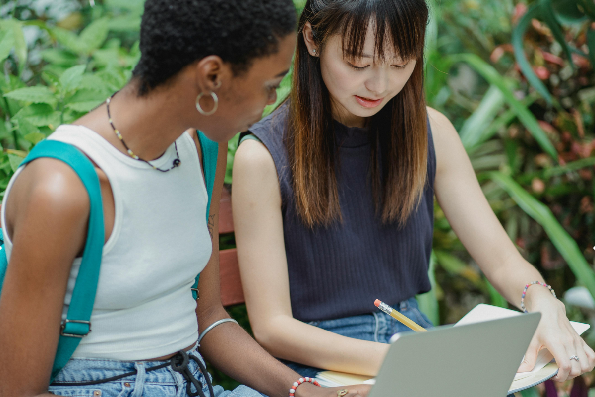 Two students work together on a laptop and review notes while seated in a leafy outdoor setting.